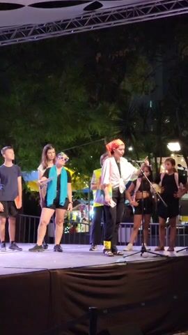 A group of seven young people, appearing to be children and teenagers, are standing on a stage at night. The stage is lit and has a dark curtain in front. The individuals are dressed in casual, colorful, and some somewhat quirky outfits. They are arranged on the stage with some facing forward and others looking towards the center. The background shows trees and some distant lights, suggesting an outdoor evening event. One person in the center, wearing a white shirt, black pants, and a red bandana, appears to be speaking or gesturing, possibly as a presenter or performer. The others seem to be listening or participating in some activity. The overall atmosphere suggests a performance or a youth event.