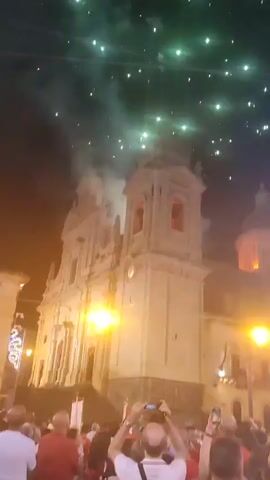 The video shows a night-time fireworks display over a building with a tall tower, possibly a church. The fireworks are bursting in green and white, with smoke visible. Below, a crowd of people watches the display. Some people are holding up their phones to record the event. Streetlights illuminate the scene. The overall atmosphere appears to be festive.