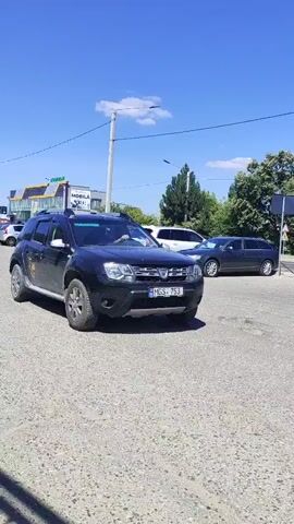 A black Dacia Duster SUV is parked on a gravel lot. The car has a license plate that reads "MGS 753". A graphic of a dial with numbers 3, 4, 5, and 6 is visible on the driver's side door. The car is dusty, with mud splatters on its lower body. Sunlight glints off the car's roof rack and side mirrors. In the background, trees and a blue sky are visible. Another dark-colored car is parked behind the Duster. A dark-colored sedan and a white SUV are parked further in the background. The scene appears to be outdoors, possibly near a road or parking area.
