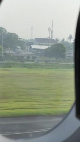 The video captures a view from an airplane window, looking out at a hazy landscape as the aircraft moves forward, likely during taxiing or takeoff. The oval frame of the window is visible, with the bottom portion showing a concrete or asphalt surface, possibly a runway or taxiway.

Directly outside the window, in the foreground, is a wide expanse of vibrant green grass, possibly rice paddies, which appears blurred due to the motion. Beyond this field, there is a distinct midground featuring a road where numerous vehicles, primarily motorcycles, are seen traveling. Several buildings, including houses with various roof types and some structures that appear more industrial or commercial, line the road. Interspersed among the buildings are numerous trees, including several tall palm trees, contributing to a tropical or subtropical feel. In the distance, a tall communication tower is briefly visible.

The background is dominated by a pale, hazy sky, which diffuses the sunlight and gives the entire scene a muted, somewhat dreamlike quality. The time of day appears to be daytime, though the haze prevents a clear indication of morning or afternoon.

The continuous sound of an aircraft engine is audible throughout the video, reinforcing the impression of being on an airplane. The rapid movement of the landscape from left to right indicates the plane is in motion. The setting, characterized by rice paddies, residential buildings, and palm trees, aligns with the provided location context of Rawa Burung, Indonesia.