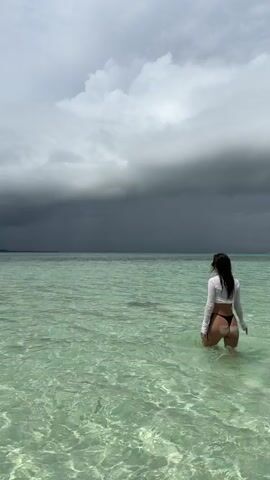 A woman is seen from behind, standing in shallow, clear, light green water. She wears a white long-sleeved top and a black bikini bottom. The sky above is overcast with dark storm clouds looming.  The horizon line is visible in the distance.  The water ripples gently.
