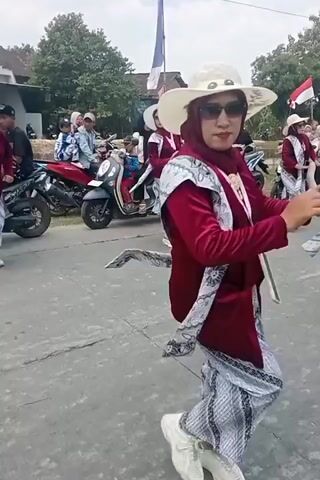The video shows a woman in a maroon outfit and a wide-brimmed hat dancing. She is wearing sunglasses and a hijab. Her outfit includes a maroon top and a patterned skirt. She is also wearing a sash draped over her shoulders. She is holding her hands up and appears to be performing a dance. In the background, there are other people, motorcycles, and buildings. The scene appears to be an outdoor event, possibly a parade or festival.