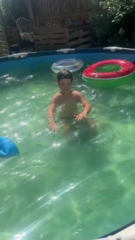 A young boy with dark hair, wearing dark swim trunks, is in a green-tinged swimming pool. He is partially submerged in the water and appears to be playing, splashing with his arms outstretched. In the background, two inflatable pool rings are floating: one with a blue and white pattern and another red and green one. A blue inflatable object is also visible in the foreground to the left. The pool is surrounded by a wooden fence and some greenery, suggesting an outdoor setting on a sunny day. The water reflects the sunlight, creating a sparkling effect.