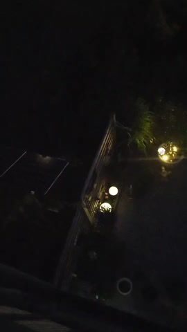 The video is a low-light, high-angle shot of an outdoor area at night. Several small, spherical lights are visible, some appearing to be garden lights. One section of the area has a structure resembling a fence or railing. There is also a dark, paved surface, possibly a patio or walkway, and some vegetation. The overall impression is of a dimly lit backyard or outdoor space.