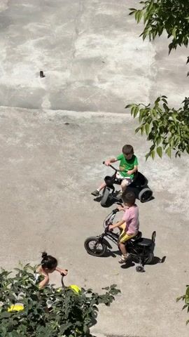 The video captures an outdoor scene on a sunny day. Two young boys are engaged in riding bicycles. The boy on the left is wearing a pink t-shirt and yellow shorts and is riding a black bicycle with training wheels. The boy on the right is wearing a green t-shirt and shorts and is riding a black tricycle with a storage bin at the back. Both boys are on a paved surface that appears to be concrete, which is somewhat uneven and shows some discoloration and cracks. There are green leafy branches from trees visible at the bottom and right edges of the frame. A third child, wearing dark clothing, is partially visible at the bottom left, appearing to be interacting with the foliage. The camera is positioned at a high angle, looking down on the scene.