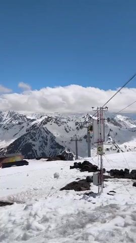 The video pans across a snowy mountain landscape under a bright blue sky with scattered white clouds. In the foreground, there is a snowy slope with dark rocks protruding. In the middle ground, a colorful building, possibly a ski lift station, sits on the snow. Further back, a ski lift with chairs is visible, ascending towards snow-covered mountains in the distance. The video continues to pan to the right, revealing more of the vast, rugged mountain range, with ski lift cables stretching across the sky. Towards the end of the clip, a few people can be seen skiing down a slope. A red net fence runs along the side of the slope. The overall scene depicts a ski resort on a sunny day with clear visibility.