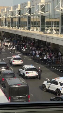 The video shows a protest taking place outside of a modern-looking building with a glass facade. There is a large group of people holding signs behind a metal barrier, and several police officers are standing on the road separating the protestors from a line of vehicles. There are several police cars and a black van. The police vehicles have the words "DIAL 911" written on them. The overall scene suggests a tense situation with a clear division between the protestors and law enforcement.