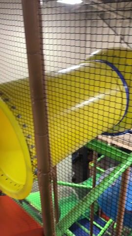 The video shows a playground structure with a yellow slide, a blue tube-like structure, and green supports, all viewed through a black net. The structure is indoors, likely in a play area or recreational facility. No individuals are clearly visible or identifiable.