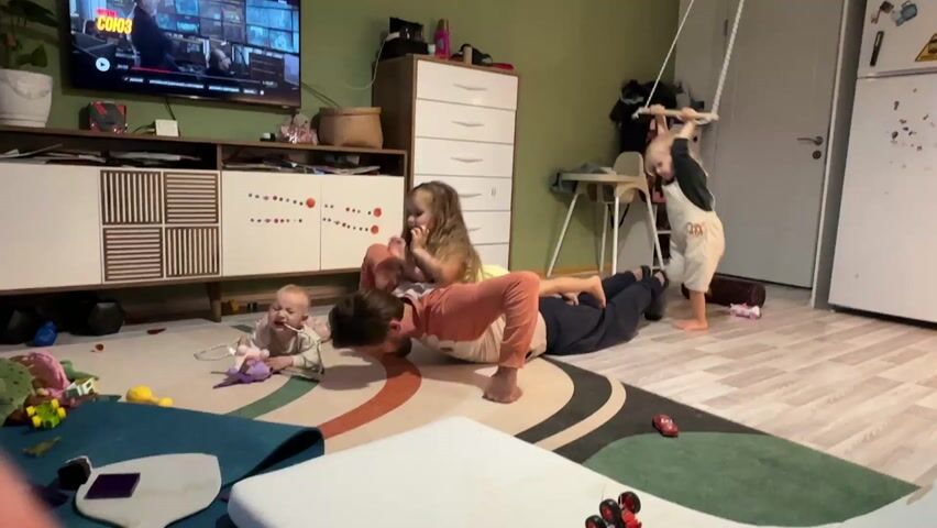 A man is doing push-ups on a rug with two children on his back, while another child swings on a suspended swing set. There is a baby crawling on the rug next to him. The room is a living room with toys scattered around, a TV playing a show, and a dresser. The overall atmosphere seems playful and energetic.