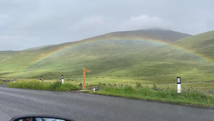 This video shows a rainbow over a green, hilly landscape. A road is in the foreground, and there are two signs by the side of the road. The sky is cloudy.