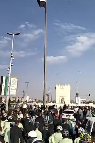 A video depicts a large group of people, mostly wearing black jackets and colorful bicycle helmets, standing around in a public square under a bright blue sky with scattered clouds. A tall flagpole with the flag of Syria waving in the wind dominates the scene. Several lampposts and a large banner displaying the Syrian flag are also visible. In the background, there's a small castle-like structure, various buildings, and traffic. The crowd appears to be gathering or waiting, but their specific activity or purpose is unclear from the footage. There are small aircraft in the sky.
