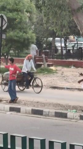 A man in a white shirt and orange head covering rides a three-wheeled bicycle laden with a large bin or cart. He is on a paved road. To his left, a young man in a red t-shirt and blue jeans stands on the sidewalk. Further left, a person in a light-colored shirt walks away from the camera, with trees and a fence in the background. To the right of the cyclist, a man in a white shirt and dark pants, wearing a turban, stands near a tree, holding a phone to his ear. His dark bag rests at the base of the tree. The environment appears to be a street with sidewalks, trees, and some greenery. The lighting suggests it is daytime.