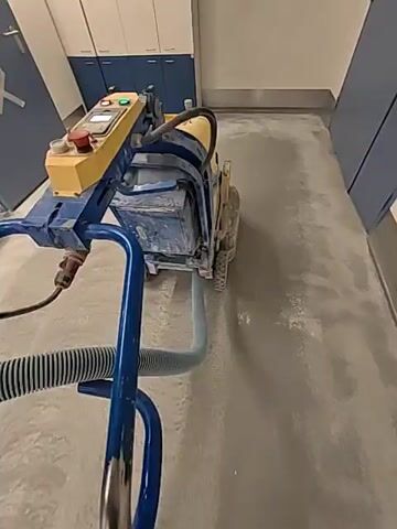 This is a first-person perspective video showing a person operating a floor grinder. The machine is yellow and blue and is connected to a vacuum hose. The floor appears to be concrete and is being ground down, creating dust. The person is wearing work boots and dark pants. The environment looks like an industrial or commercial space with cabinets and doors in the background. The video captures the process of floor preparation or renovation.