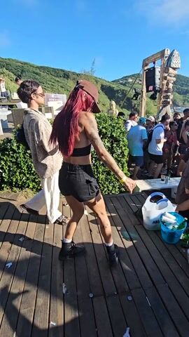 The video shows a woman dancing on a wooden deck at an outdoor event. She has red hair, tattoos on her arms, and wears a black leather-like top, black shorts, and black boots. A brown baseball cap shadows her face. She is dancing with arm and leg movements in time to the music.

In the background, there are people mingling, foliage, a wooden structure that appears to be part of a stage, and green hills. There are also items like a white plastic bag, an ice bucket with bottles, and other miscellaneous objects scattered around the deck. The sky is clear and blue. There are other people standing behind and to the side of the woman, including one person with sunglasses and light-colored clothing.