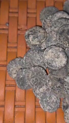 A close-up, overhead shot displays a pile of ancient, weathered coins. The coins are dark and appear to be made of a metallic substance, possibly bronze or copper, with some showing signs of patina and corrosion. They are irregularly shaped and have visible markings or designs on their surfaces, though these details are obscured by age and wear. The coins are clustered together, partially overlapping, on a textured surface that resembles woven bamboo or straw, which is a reddish-brown color. The lighting is dim, casting subtle shadows on the coins and highlighting their rough textures.