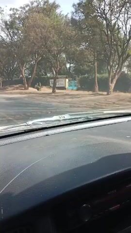 The video shows a view from inside a car driving down a road. The road is flanked by trees and a school sign for Wogo Dei Comprehensive School. The car passes a white Toyota Hiace van with the license plate KDC 660B. The sky is partly cloudy.