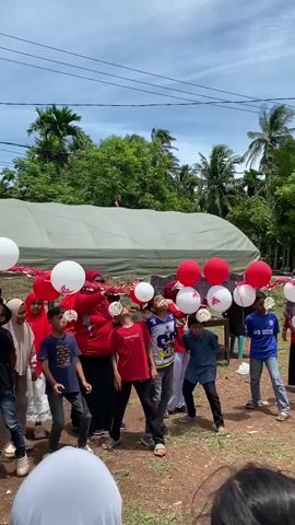 The video shows a group of children and adults gathered outdoors, possibly at a celebration or festival. In the foreground, several children are participating in a game where they attempt to eat crackers or biscuits suspended from strings. Red and white balloons are visible, hanging from a string above the children, some with white circular decorations on them. The children are looking up, with crackers in their mouths, and appear to be actively engaged in the challenge. Some adults, dressed in red clothing, are supervising the game. The background features a large tent or canopy, trees, and a bright sky, suggesting a sunny day. The ground appears to be dirt or gravel.