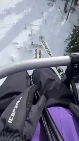A person wearing a purple ski jacket and a helmet with blue tinted goggles is recording a video from a ski lift. The person is facing the camera. The chairlift is going up a snow covered mountain with trees. The person's skis are visible below.