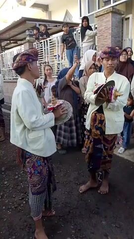 A group of young men wearing traditional clothing are playing instruments in what appears to be a street or pathway. The men are barefoot and are wearing sarongs and white shirts, with some wearing headscarves or turbans. They are holding small cymbals or hand drums and appear to be playing them in unison. A crowd of onlookers, including women and children, are standing to the side, some of whom are recording the performance on their phones. The background shows houses and some vegetation. The lighting suggests it is daytime.