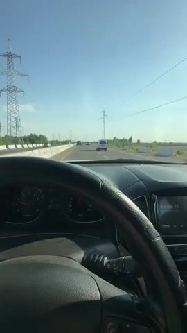 The video captures the driver's perspective from inside a car on a highway. The steering wheel and dashboard are prominent in the foreground, with the speedometer showing a speed of approximately 100 km/h. Multiple vehicles are visible on the highway ahead, including cars and a white van. Power lines and utility poles run parallel to the road, indicating an open, rural or semi-rural environment. The sky is clear and blue, suggesting a sunny day. The road appears to be a multi-lane highway with a concrete barrier on the left side. The overall scene depicts a typical driving experience on a highway.