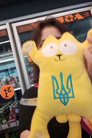 A woman is holding a yellow stuffed cat with the Ukrainian trident symbol on its chest. She is smiling and looking at the camera. The background appears to be a store aisle with refrigerators and shelves stocked with products. There are also some orange circular signs with black text. The woman has brown hair and is wearing a black t-shirt with a pink strap over her shoulder.