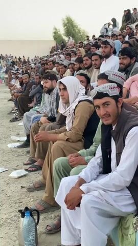 The video shows a crowd of men, mostly young adults and middle-aged, sitting on tiered bleachers. They are dressed in traditional clothing, including shalwar kameez and vests, with many wearing kufi caps. The atmosphere appears to be an outdoor gathering, possibly a sporting event or a public assembly, as suggested by the bleacher seating and the focused attention of the individuals. Some people are looking towards the left, out of frame, while others are looking towards the camera. There are a few plastic bottles and containers scattered on the ground in front of the bleachers. The environment appears to be dusty and arid, with a neutral-colored background that suggests an outdoor, possibly rural, setting. The lighting indicates it is daytime, possibly late afternoon.