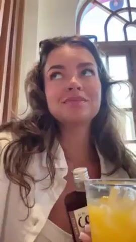 A woman with long curly brown hair and sunglasses on her head is pouring cold brew from a small glass bottle into a glass of orange juice. She is wearing a white top and sitting in what appears to be a restaurant, evidenced by the wooden paneling and architectural details of the window behind her. The bottle is labeled "Cold Brew." The action focuses on the pouring motion and the mixing of the two liquids.