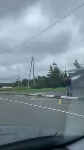 The video shows a view from inside a vehicle driving on a road under a cloudy sky. There are power lines and utility poles visible along the side of the road. In the first few seconds, a person with a bag is seen walking near the side of the road, and a white car is parked in a grassy area. Road markings, including arrows indicating traffic direction, are visible on the pavement. A sign with Cyrillic text and arrows is also present. Several vehicles, including a dark van and a black SUV, are seen driving on the road. The scenery includes trees and fields.