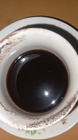 A close-up shot shows a cup of coffee on a saucer. The coffee is dark brown and appears to have some sediment at the bottom. The cup and saucer are white with a floral pattern. The saucer is slightly chipped. The background is a wooden table.