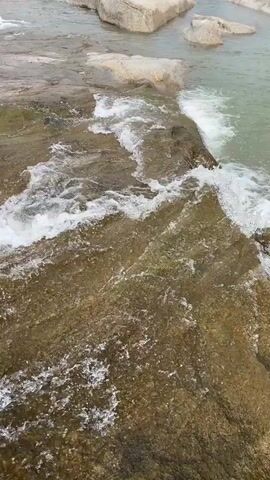 The video shows a stream of water flowing over rocks. The water is mostly clear, with some areas of white foam where it rushes over the rocks. The rocks are a mixture of light gray and brown. The camera angle is from above, looking down at the water.
