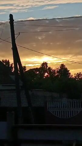The video shows a sunset with trees in the foreground. The sun is visible through the trees, casting a warm glow. There are clouds in the sky. There are also power lines and a utility pole visible. In the foreground, there is a brick structure and a fence. The video is shaky, suggesting it was filmed handheld.