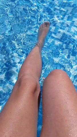 The video shows a first-person perspective of someone's legs and feet submerged in a swimming pool. The water is clear and blue, with sunlight creating shimmering patterns on the pool floor. The person is moving their feet and legs through the water, causing ripples and splashes. The camera angle is looking down at the legs and feet, with the water filling most of the frame.