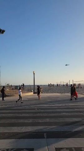 A helicopter flies in the sky over a beach boardwalk. People are walking on the boardwalk and on the beach. There are volleyball nets on the beach. The sun is setting or rising.