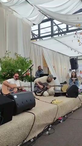 A group of musicians are performing on a stage under a white tent. Two men are playing acoustic guitars, one standing and one seated. A third man is playing a keyboard behind them. A young woman is seated on the right, singing into a microphone. There are speakers on the stage, and the floor is covered with a beige rug. The background is decorated with white drapes and some greenery.