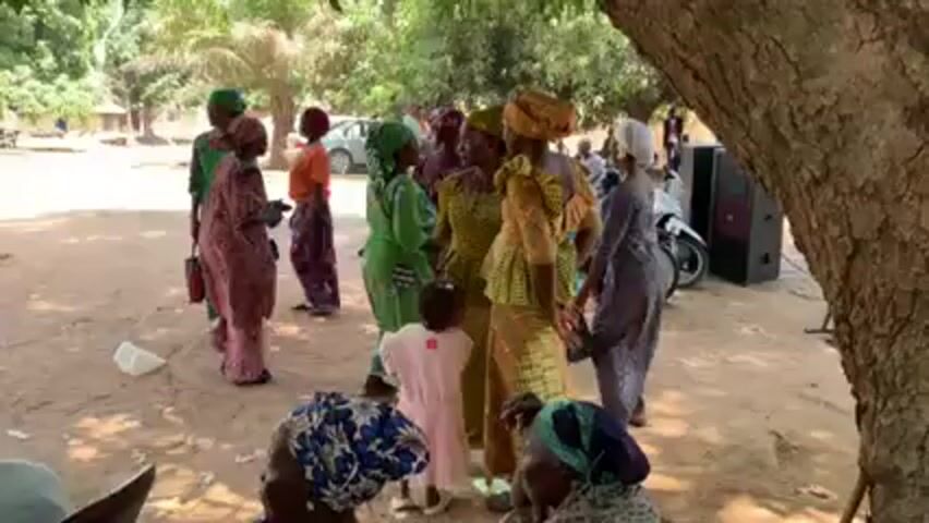 The video shows a gathering of women in an outdoor setting, possibly a village. Most of the women are wearing traditional African dresses in various vibrant colors and patterns, and they are adorned with headwraps. They are standing or milling about on a sandy ground under the shade of trees. A young child in a pink dress stands near the center of the group. There's a parked car in the background, a motorcycle, and some speakers set up, suggesting the possibility of music or an event. Some women are in conversation, while others seem to be greeting or acknowledging each other. The atmosphere appears lively and social.