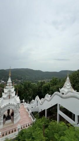 The video shows a view from a height looking down on white tiered structures with golden spires, resembling a temple or pagoda, set against a backdrop of lush green mountains under a cloudy sky. A white, serpentine railing with a decorative pattern, possibly depicting scales, lines a set of stairs. The foreground is dominated by the vibrant green foliage of trees and bushes. On the platform of the temple, two figures, likely visitors, are visible near an entrance. The scene is peaceful and serene, suggesting a temple complex located in a natural, elevated setting.