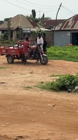 In Jawu, Nigeria, three young men are gathered around a red three-wheeled motorcycle-like vehicle with a cargo bed. One man, wearing a red t-shirt and shorts, stands by the vehicle's front wheel, appearing to adjust or interact with it. Another man, dressed in a white robe, sits on the motorcycle, facing away from the camera. A third person, wearing dark clothing, stands behind the man on the motorcycle.

The scene unfolds on a dirt road in front of a row of buildings with corrugated metal roofs, indicative of a rural or semi-urban setting. The sky is overcast, suggesting a cloudy day, possibly humid. Green foliage grows around the buildings and in the foreground. The overall atmosphere seems casual, with the men engaged in some activity involving the vehicle.

Visible text on the red cargo bed includes "GP" and "MOBILE." A sign on one of the buildings reads "POS," likely indicating a Point of Sale establishment. The houses appear modest, with one having a sign that is difficult to read but seems to be for a business. The ground is dry and dusty, consistent with the unpaved road. The time of day appears to be daytime, but the exact hour is unclear due to the overcast conditions. The interaction between the individuals suggests a shared task or perhaps preparation for travel or delivery.