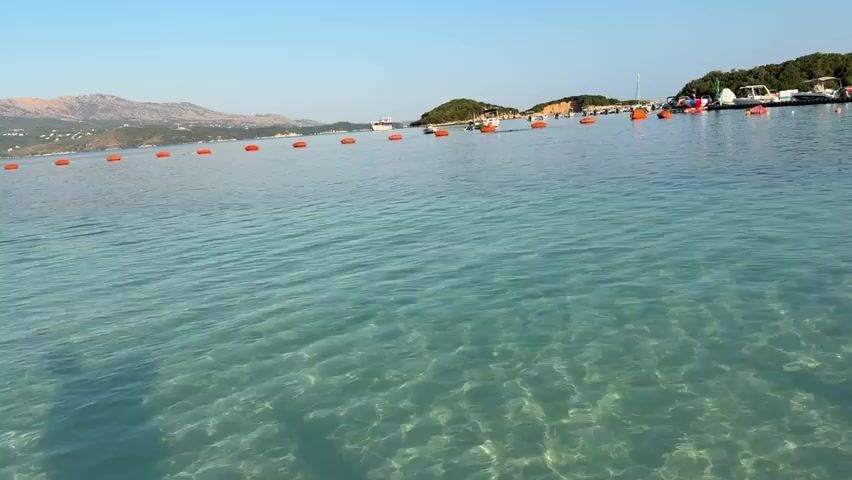 The video shows a calm, clear turquoise sea with small waves. The seabed is visible through the clear water. In the distance, there is a shoreline with green hills and some trees. There are several small boats and a pier with some structures and people on it. A line of orange buoys stretches across the water, marking a boundary. The sky is clear and blue, indicating a sunny day. The camera appears to be moving slightly, giving a sense of gentle motion on the water.