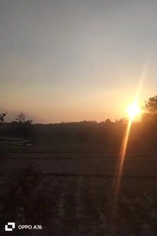 The video shows a sunset over a rural landscape. The sun is low in the sky, casting a warm glow over the scene. The foreground is a field of dark, tilled earth, possibly a farm field. In the middle ground, there are trees and vegetation, with a cell tower visible behind them on the right. The sky is a gradient of pale orange and yellow near the horizon, transitioning to a lighter gray at the top of the frame. The sun itself is a bright, lens-flared orb positioned slightly to the right of the center of the image. The overall impression is one of peace and natural beauty. The video is stamped with "OPPO A76" in the bottom left corner.
