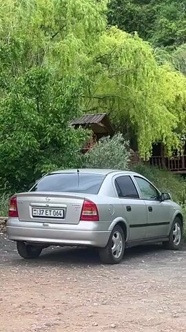 A silver four-door sedan is parked on a dirt road in the foreground, facing right. To the right, nestled into a green, tree-covered hillside, is a rustic wooden structure with a sloped roof and an open balcony. Several people are visible on the balcony; one person in a light-colored top stands at the railing. The background consists of a steep, verdant hillside. The camera slowly pans to the right, revealing more of the green landscape and the wooden structure.