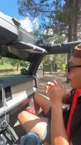 A man is driving a 2021-2023 Ford Bronco, based on the vehicle's dash. The top of the Bronco is removed. The man is wearing glasses, a hat, a black shirt with a red strap, and shorts, and has a water bottle near his legs. The Bronco is on a dirt road surrounded by trees and grass. The sky is blue with some clouds. The man has his hands cupped together in front of his mouth.