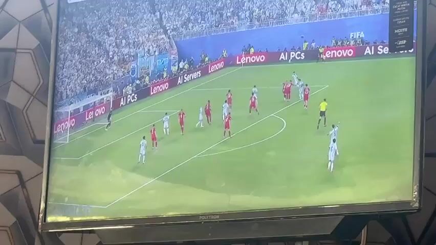 The video shows a close-up of a television screen displaying a soccer match. The game is being played on a bright green field with white markings. Several players in red and white uniforms are visible on the field. A referee in a yellow shirt is also present. The background shows a stadium filled with spectators, and advertisements are displayed around the field. The image quality is slightly blurry.