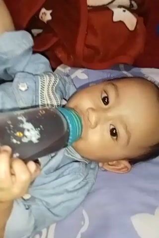 Here is a forensic description of the video:

A baby wearing a blue denim shirt is lying on their back on a bed with blue patterned sheets. The baby is holding a clear plastic bottle filled with a dark liquid and drinking from it. The bottle has a light blue nipple and cap. A red blanket with a white design is visible in the upper left corner of the frame. The baby is looking up and to the right, then looks towards the camera. The baby has dark hair and brown eyes. The shot is static, with a high-angle view.