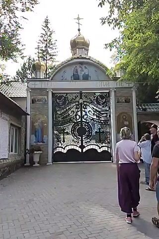 A group of four people walks towards a large ornate gate leading to a monastery. The gate is black with intricate metalwork and features crosses and decorative swirls. Above the gate is an archway with religious iconography and a golden dome topped with a cross. To the left of the gate is a stone building with a tiled roof. The ground in front of the gate is paved with bricks. Trees and foliage surround the entrance. The people, two women and two men, are walking away from the camera towards the gate. The woman in the foreground is wearing a pink top and purple pants. The man next to her is wearing a black t-shirt and denim shorts. Another woman in a white top and jeans is walking behind them.