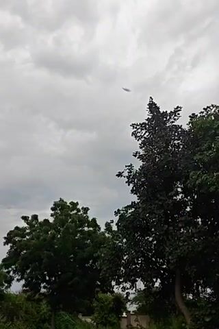The video shows a tilted shot of a cloudy sky with trees. A white building with dark windows appears on the left side of the screen.