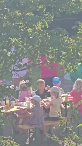 A group of children are gathered around a picnic table in a sunlit outdoor setting, possibly a campground. They are eating and interacting with each other. The scene is partially obscured by lush green foliage, and in the background, several RVs and trees are visible under a bright, clear sky.