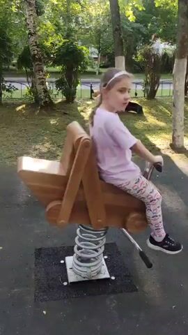A young girl with blonde hair in a ponytail, wearing a pink t-shirt and patterned leggings, sits on a wooden spring rider designed to look like a stroller. She is holding onto the handlebars and bouncing. The spring rider is on a black rubber safety surface in a park. Behind her, there are trees and a white fence. In the background, there appear to be two lion statues visible.