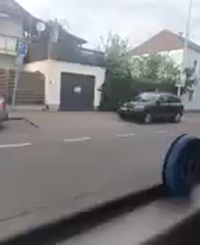 The video shows a street scene in Ginsheim-Gustavsburg, Germany, on an overcast day. In the background, there are several buildings, including a light-colored house with a dark garage door and a separate white building. Greenery, possibly bushes or trees, is visible between the buildings. A black car is parked on the side of the road, facing towards the right of the frame. White dashed lines are painted on the asphalt road surface. From the left, a silver car drives past the parked black car and exits the frame to the right. A blue object is partially visible in the bottom right corner of the foreground, suggesting the recording is made from inside a vehicle or structure. A blue "P" sign, indicating parking, is partially visible on the left side of the frame, near the light-colored house.