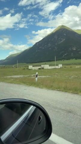 A car is driving down a road with a mountain landscape. A side mirror of the car shows the reflection of the mountain and sky. In the distance, a herd of sheep is grazing in a green field. The sky is blue with white clouds.