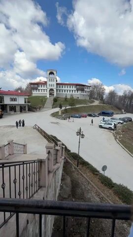The video shows an outdoor view of what appears to be a religious complex. In the primary view, there is a large, white building with a red roof and a central tower, situated on a small hill. A pathway leads up to it, and there is a parking area nearby with several cars and a blue bus. People are visible, walking around in the plaza. The sky is blue with scattered white clouds.

The camera then pans left, showing more of the complex, including a smaller building with a red roof. A truck is parked nearby.  

Next, the camera shifts to show a stone terrace or balcony area, with a metal railing and a view of a forest in the distance. 

Finally, the camera focuses on a building with arched doorways, brick accents, and a religious mosaic on the wall. Several people are entering the building.